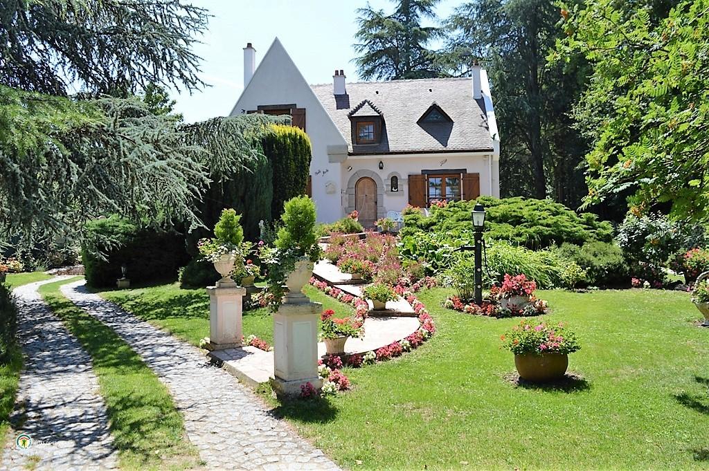 A charming house is framed by lush greenery and vibrant flower beds, creating a peaceful outdoor ambiance. A stone pathway leads to the entrance, flanked by ornamental planters filled with colorful blooms. Tall trees provide shade, enhancing the serene setting.