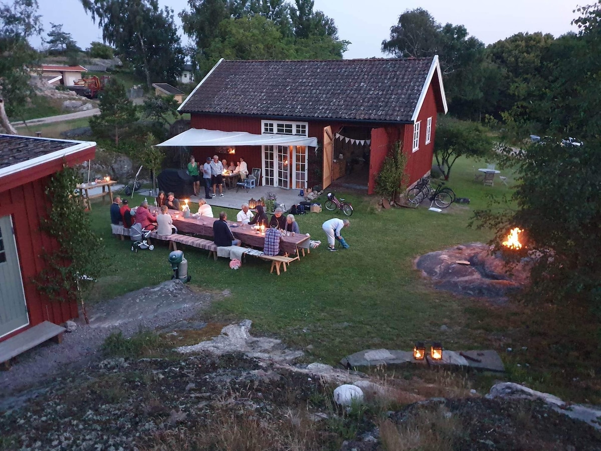 An outdoor gathering is depicted in a lush green yard, featuring a long picnic table set with candles. Nearby, a fire pit glows softly as guests enjoy the evening. The red structure in the background is complemented by bicycles and a cozy atmosphere.