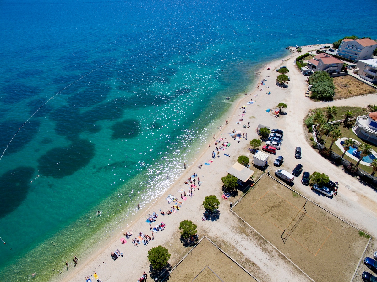 Aerial view captures a serene beachfront with soft sandy shores and crystal-clear waters. Sunbathers can be seen scattered along the beach, while vehicles are parked nearby. Volleyball courts are present, contributing to the beachfront activity, surrounded by lush greenery and coastal properties.