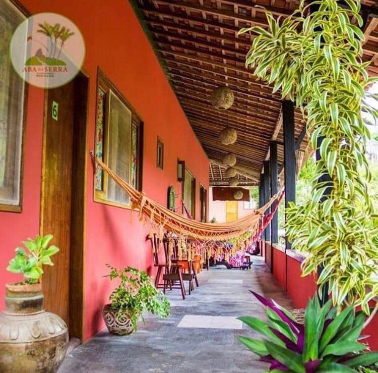 A vibrant hallway is highlighted by colorful hammocks and wooden chairs lined along the walls. Lush green plants are positioned strategically, adding a natural touch. The traditional architecture features a wooden ceiling and terracotta-red walls, creating a welcoming and relaxed atmosphere.