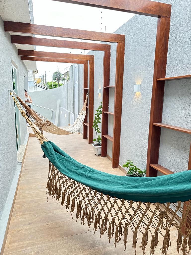 A serene outdoor corridor features two hammocks suspended between wooden beams. The space is lined with shelves and small potted plants, providing a calm atmosphere. Soft natural light filters through the area, offering a peaceful setting for relaxation.