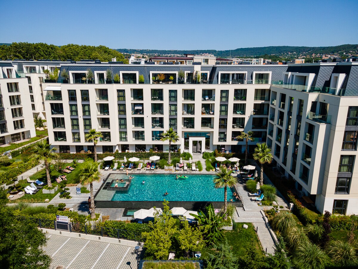 The image captures a modern residential complex featuring a large swimming pool at its center, surrounded by palm trees and sun loungers. Clear blue skies accentuate the well-maintained landscape, which includes green plants and organized seating areas.