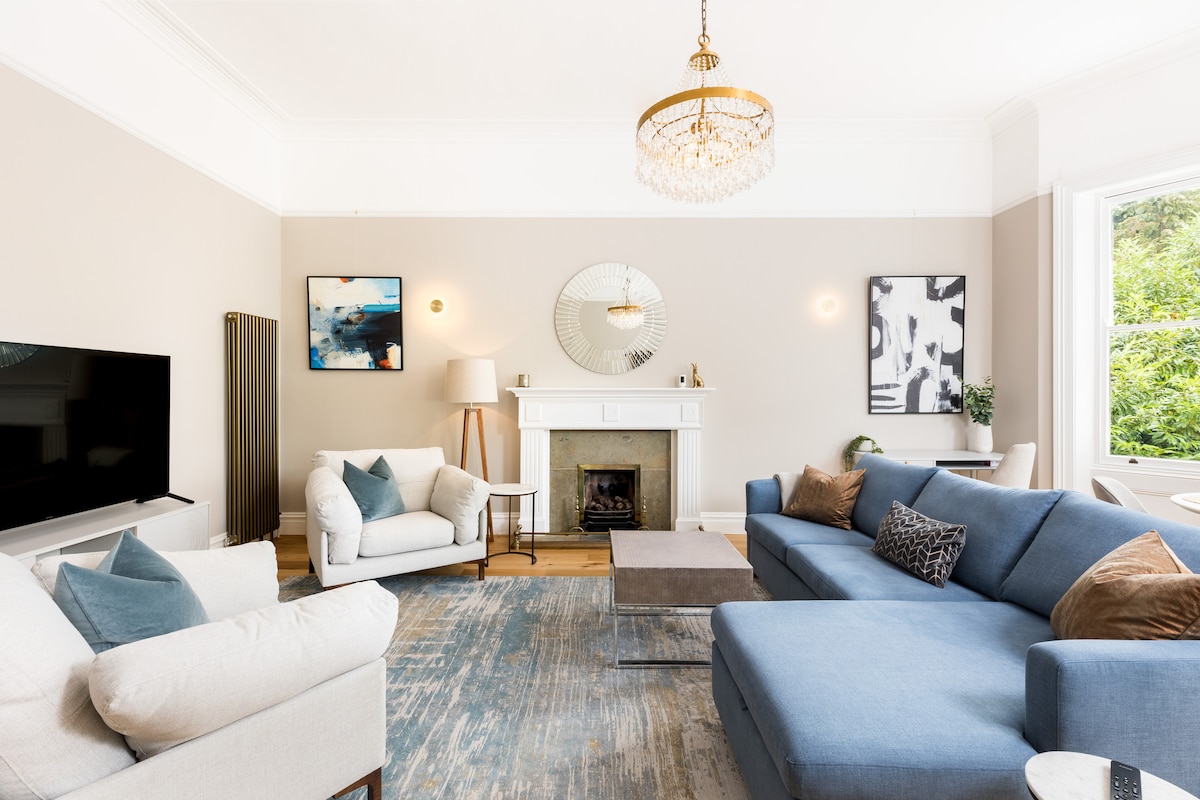 An open-plan living area is highlighted by a large light fixture and a decorative fireplace. A blue sofa and an accent chair are arranged thoughtfully around a coffee table. Natural light filters in through the bay window, illuminating the soft color palette and modern decor.