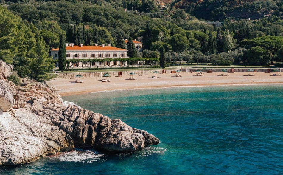 A serene beach scene is captured, with soft sandy shores curving along a calm, turquoise sea. Lush greenery lines the background, and a large building is visible at the edge of the beach, surrounded by umbrellas and sunbeds for relaxation.