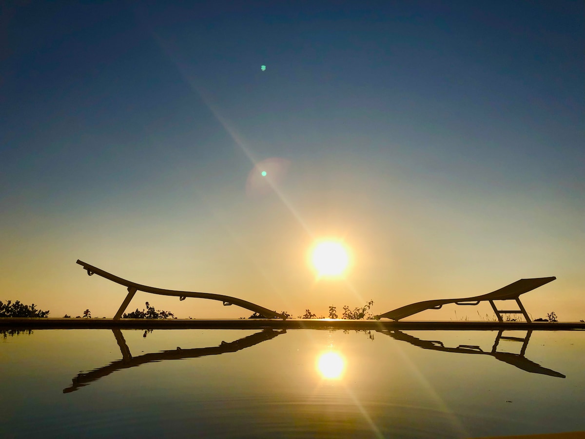 Two lounge chairs are positioned beside a calm body of water, reflecting the warm colors of a sunset. The silhouette of the chairs contrasts against the vibrant sky, with the sun setting in the background, creating a serene atmosphere.