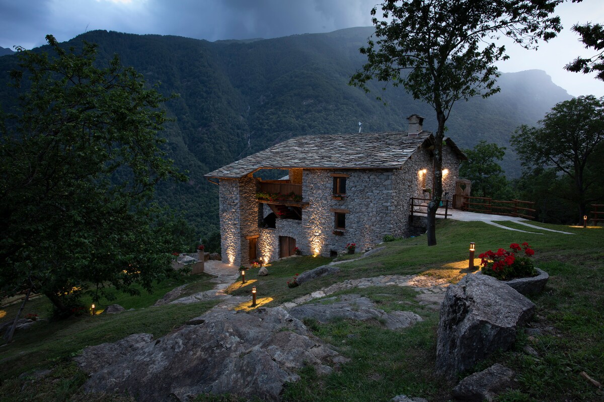 A stone house with a sloped roof sits against a mountain backdrop, surrounded by greenery. Soft lighting highlights the structure's features and pathway, while potted flowers add a touch of color to the landscape. The setting conveys a serene atmosphere amidst nature.