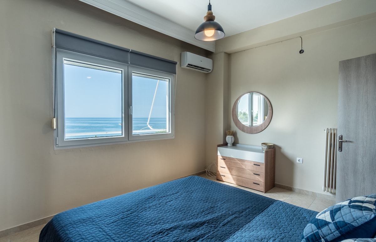 A bright sleeping area features a spacious bed with a textured blue coverlet. A round mirror is positioned above a minimalist dresser, and large windows allow natural light to flood the room, revealing a view of the ocean and a serene blue sky.