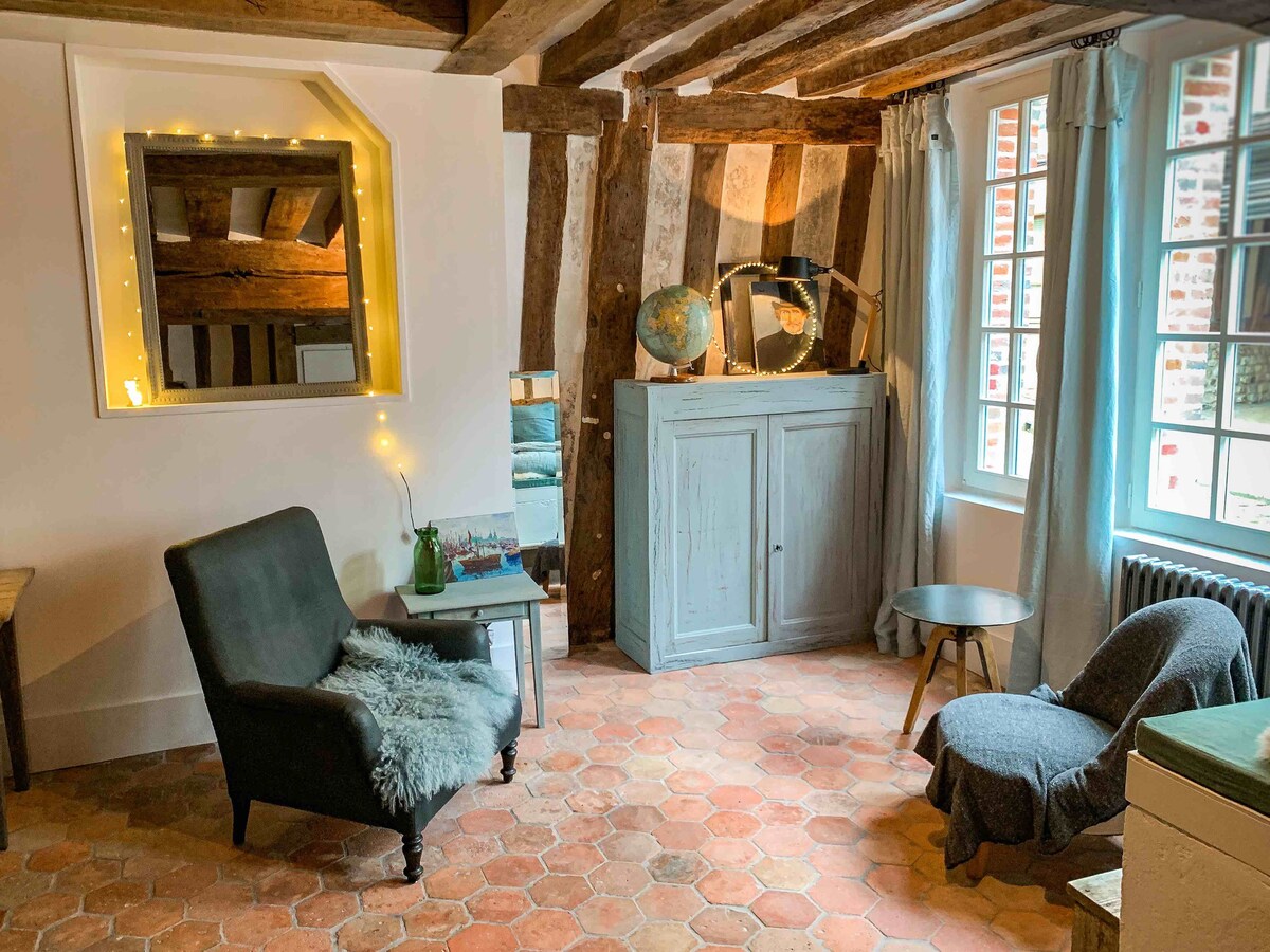 A cozy seating area features a comfortable chair and a small side table on a terracotta tiled floor. Exposed wooden beams are visible on the ceiling, while a large window allows natural light to illuminate the space. Artwork and a globe add character to the room.