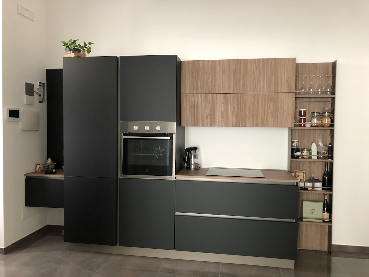 A modern kitchen is organized with sleek black cabinetry and wooden accents. Essential appliances, including an oven and kettle, are visible. Glassware and pantry items are neatly arranged on open shelves, creating a functional and minimalist design.