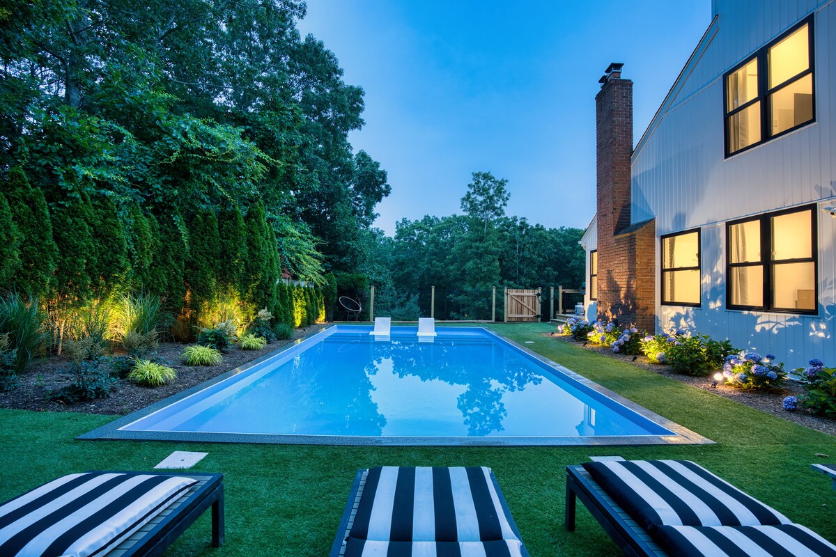 A modern swimming pool is nestled in a landscaped backyard, surrounded by greenery and colorful flowers. The water reflects the evening sky, while lounge chairs with striped cushions are placed nearby. Gentle lighting enhances the serene ambiance of the outdoor space.