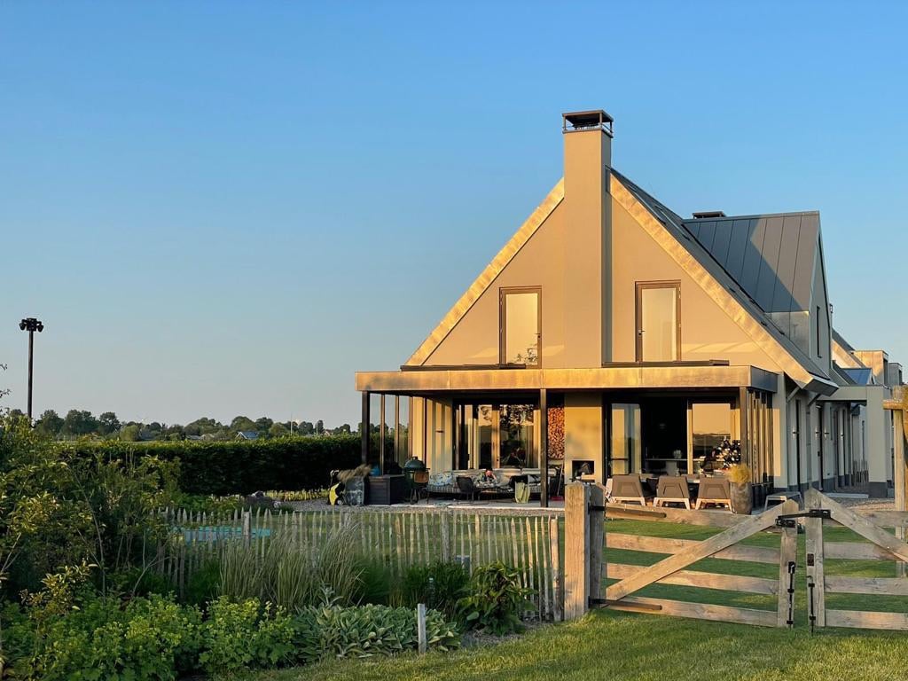 A modern villa is showcased with a distinctive triangular roof. Large windows provide a view of the outdoor seating area. A well-maintained garden with greenery surrounds the property, and a wooden fence frames the entrance, accentuating the tranquil rural setting.