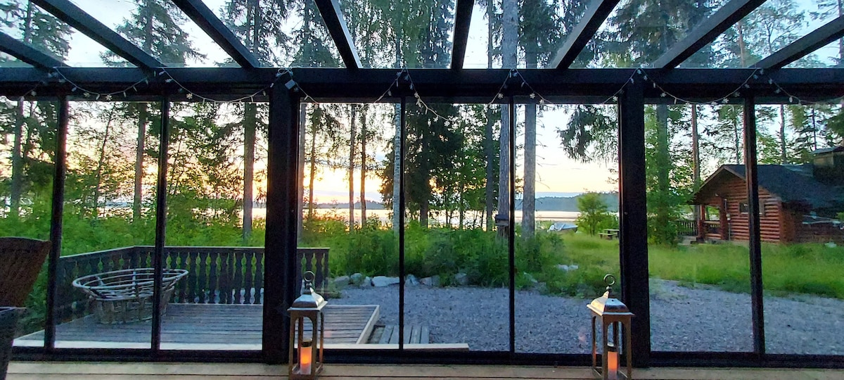 A large glass terrace offers views of the serene lake and surrounding greenery. Lanterns on the terrace provide soft lighting, while trees frame the scene. The sunset casts warm tones across the landscape, emphasizing the tranquil outdoor environment.