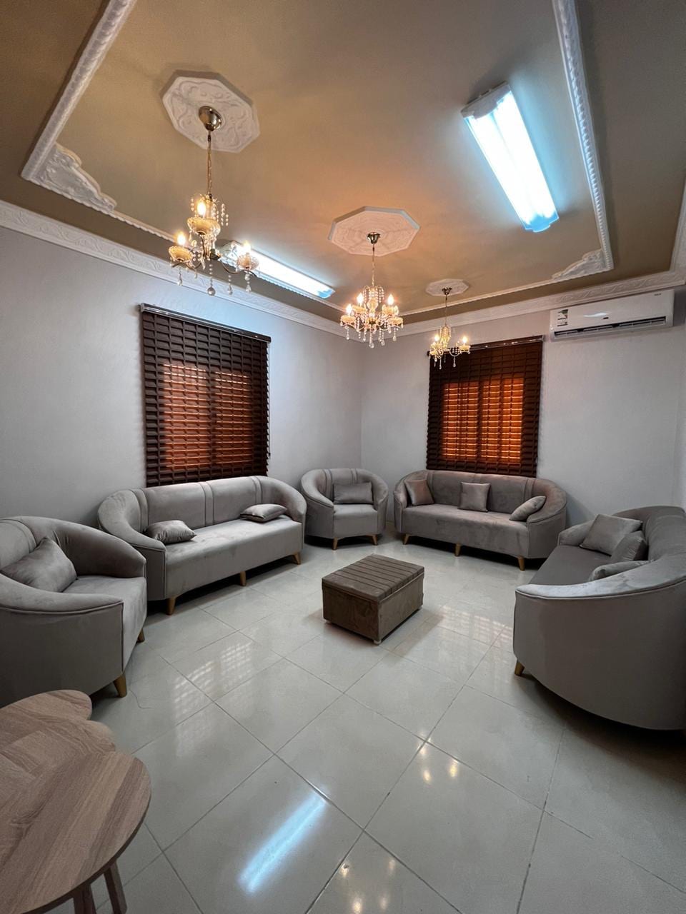 The living room features a cozy seating arrangement with curved, plush sofas and a central coffee table. Two windows are adorned with wooden blinds, allowing natural light to filter in. Elegant chandeliers hang from the ceiling, providing ambient lighting.