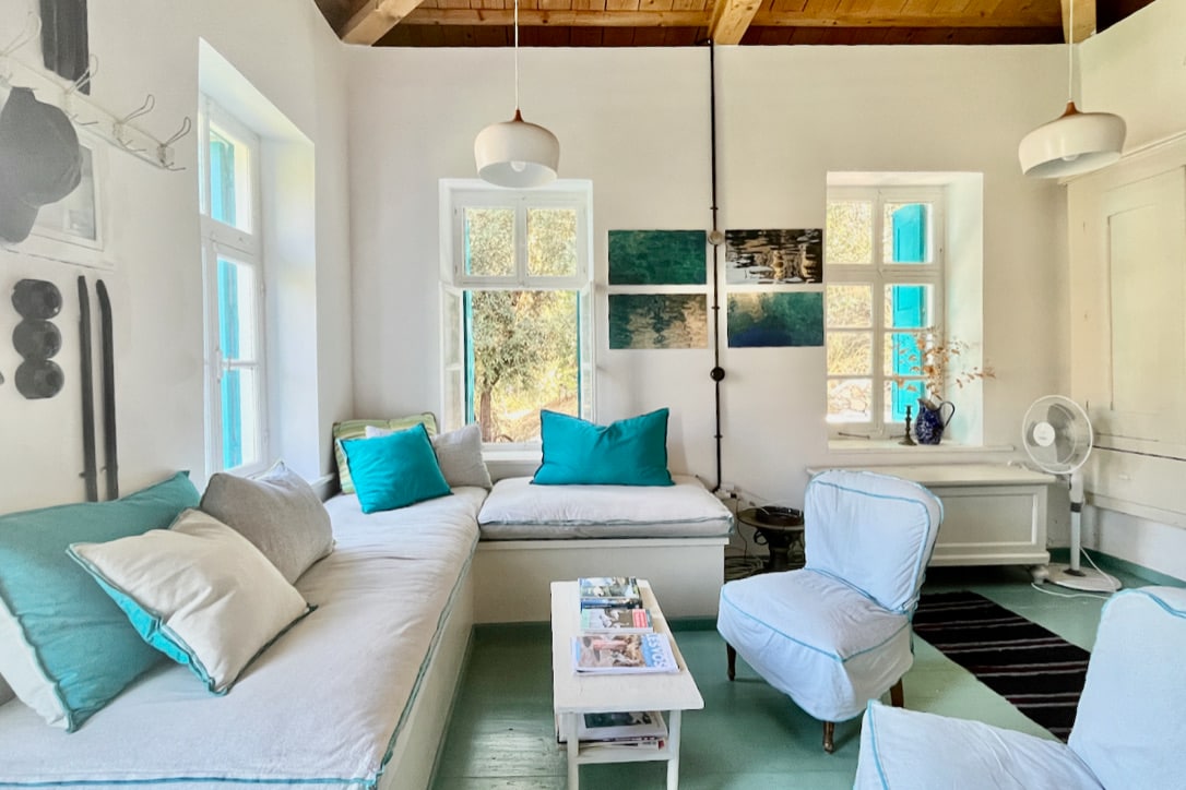 A bright living space is showcased with a sunlit corner sofa adorned with turquoise and neutral cushions. Soft light filters through multiple windows, highlighting the minimalist decor. A small white coffee table rests on a light wooden floor, complemented by a cozy armchair.