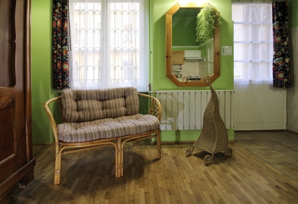 A cozy seating area is presented with a striped couch and a decorative wall mirror. Green walls enhance the brightness of the space, while natural light filters through the double windows adorned with patterned curtains. The wooden floor adds warmth to the room.