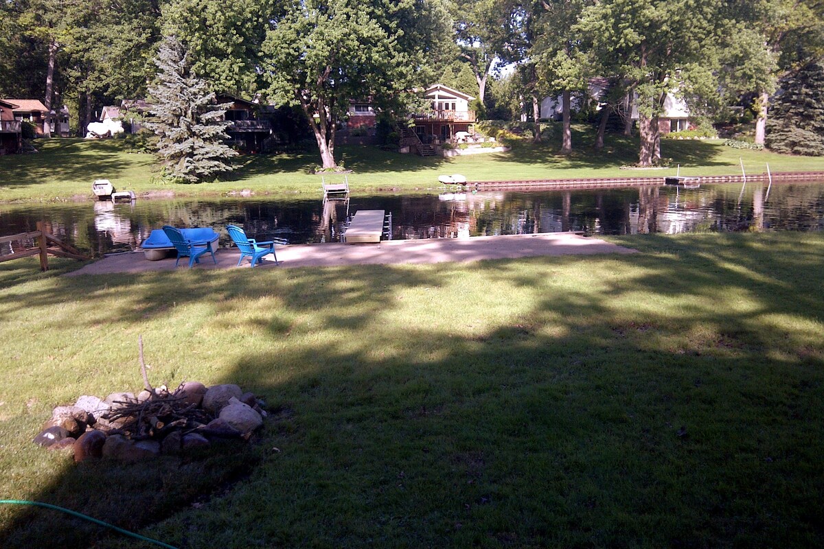 A grassy area features a sandy space extending to the water's edge, where a calm lake reflects the surrounding trees. Two blue lounge chairs are positioned nearby, and a wooden dock stretches into the water. A fire pit made of stones is located in the foreground.