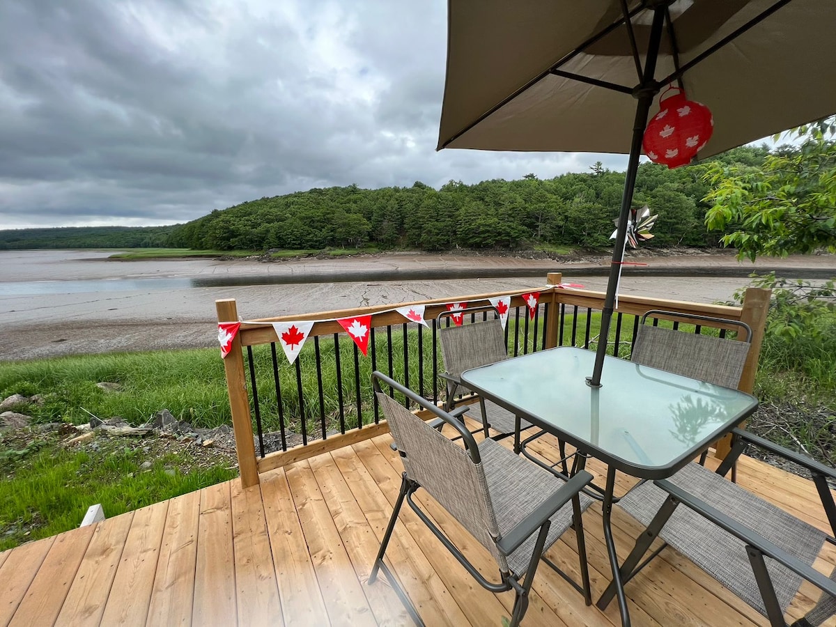 A wooden deck features a glass-top table surrounded by four chairs, complemented by an umbrella. Red and white decorative banners hang along the railing. The view includes a riverbank lined with greenery and a cloudy sky, suggesting a tranquil outdoor space.