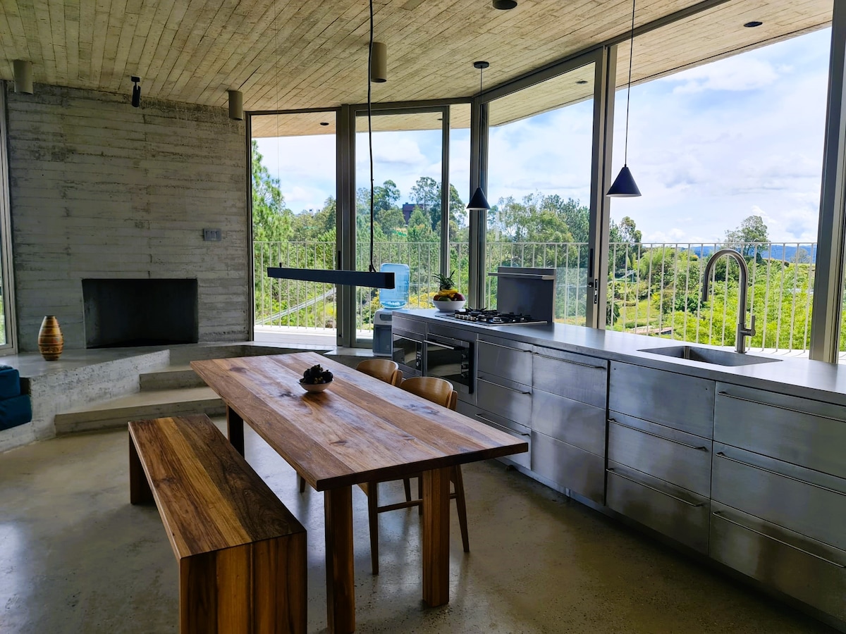 An open kitchen and dining area feature large windows that provide expansive views of the surrounding greenery. A substantial wooden dining table is accompanied by sleek chairs. Stainless steel cabinetry and a modern kitchen setup enhance the contemporary design, while a fireplace is positioned against the textured concrete wall.