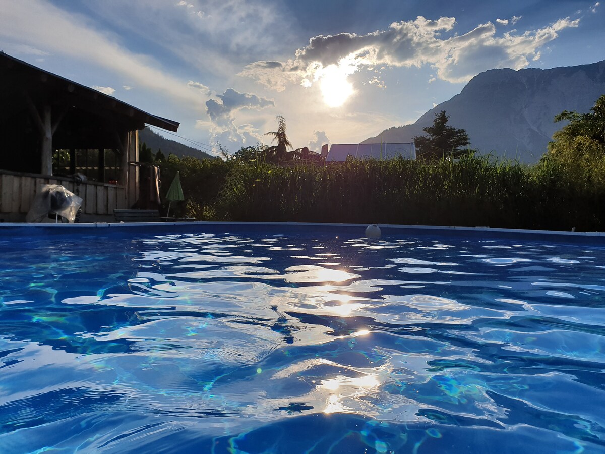The swimming pool reflects the vibrant blue sky, with sunlight shimmering on its surface. Surrounding greenery creates a natural backdrop, while the distant mountains are partially visible, enhancing the calm setting. A wooden structure is present nearby, adding to the outdoor ambiance.