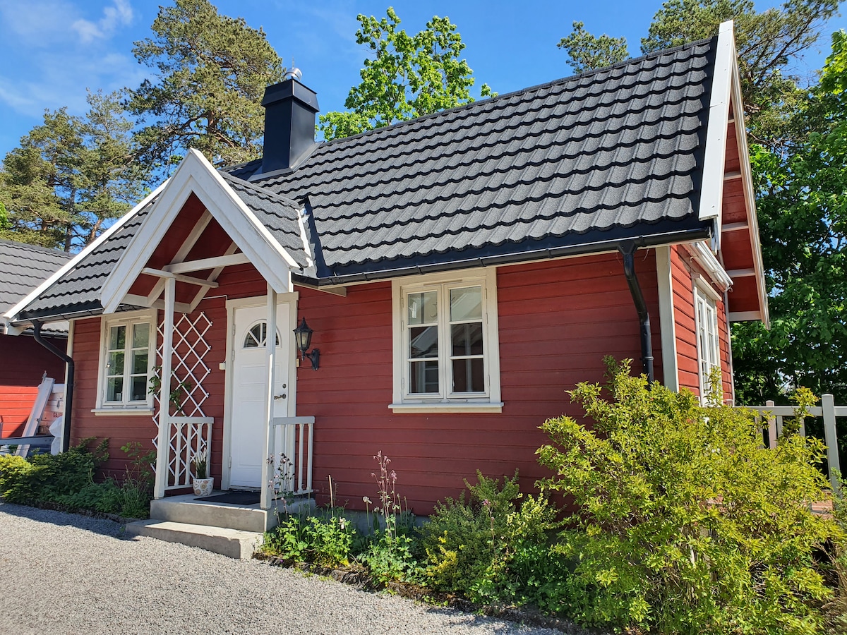 A charming red cottage with a black shingle roof is set amidst trees and greenery. White window frames contrast with the exterior, and a small porch is visible at the entrance, enhancing the inviting appearance of the home.