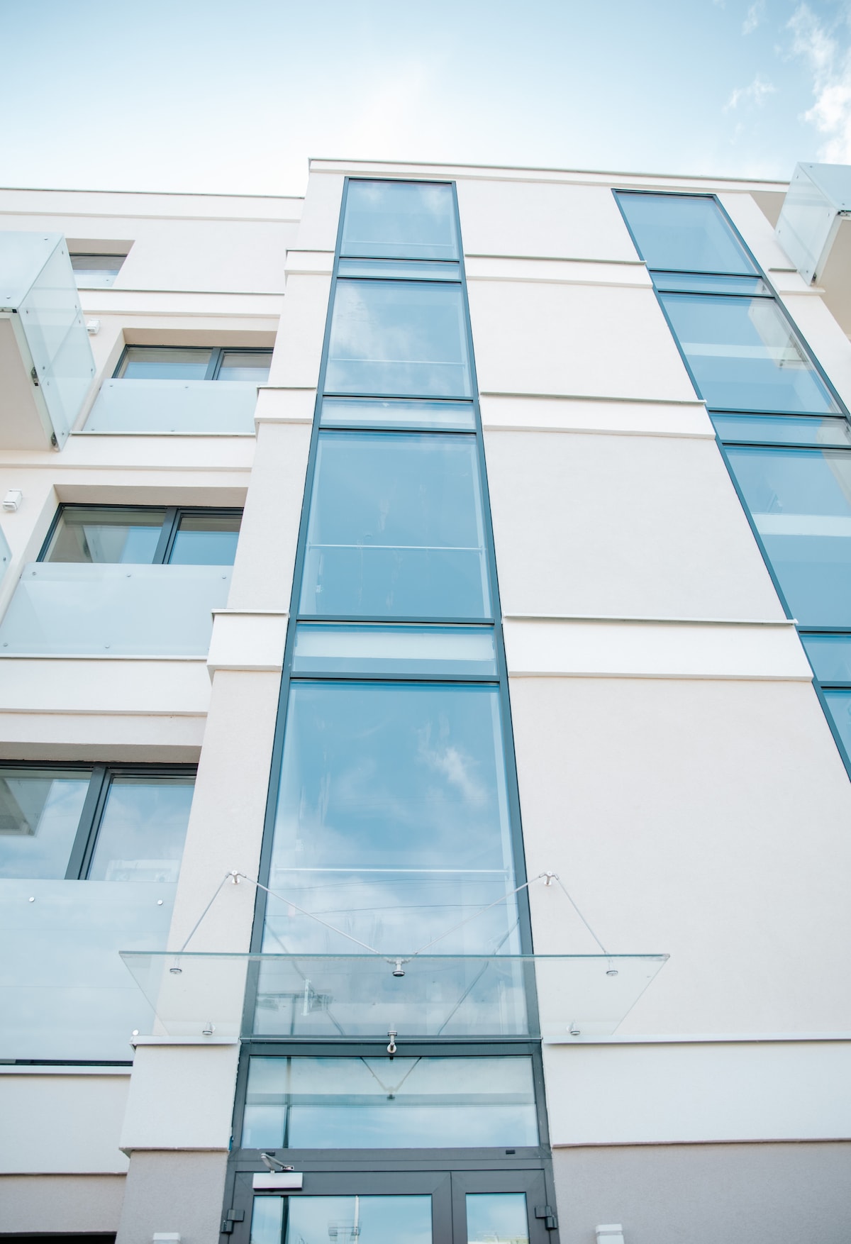 The exterior of a modern building is featured, showcasing an impressive glass facade and a sharp architectural design. A transparent canopy extends from the entrance, supported by sleek metal fixtures. Clear blue skies and a few clouds provide a serene backdrop.
