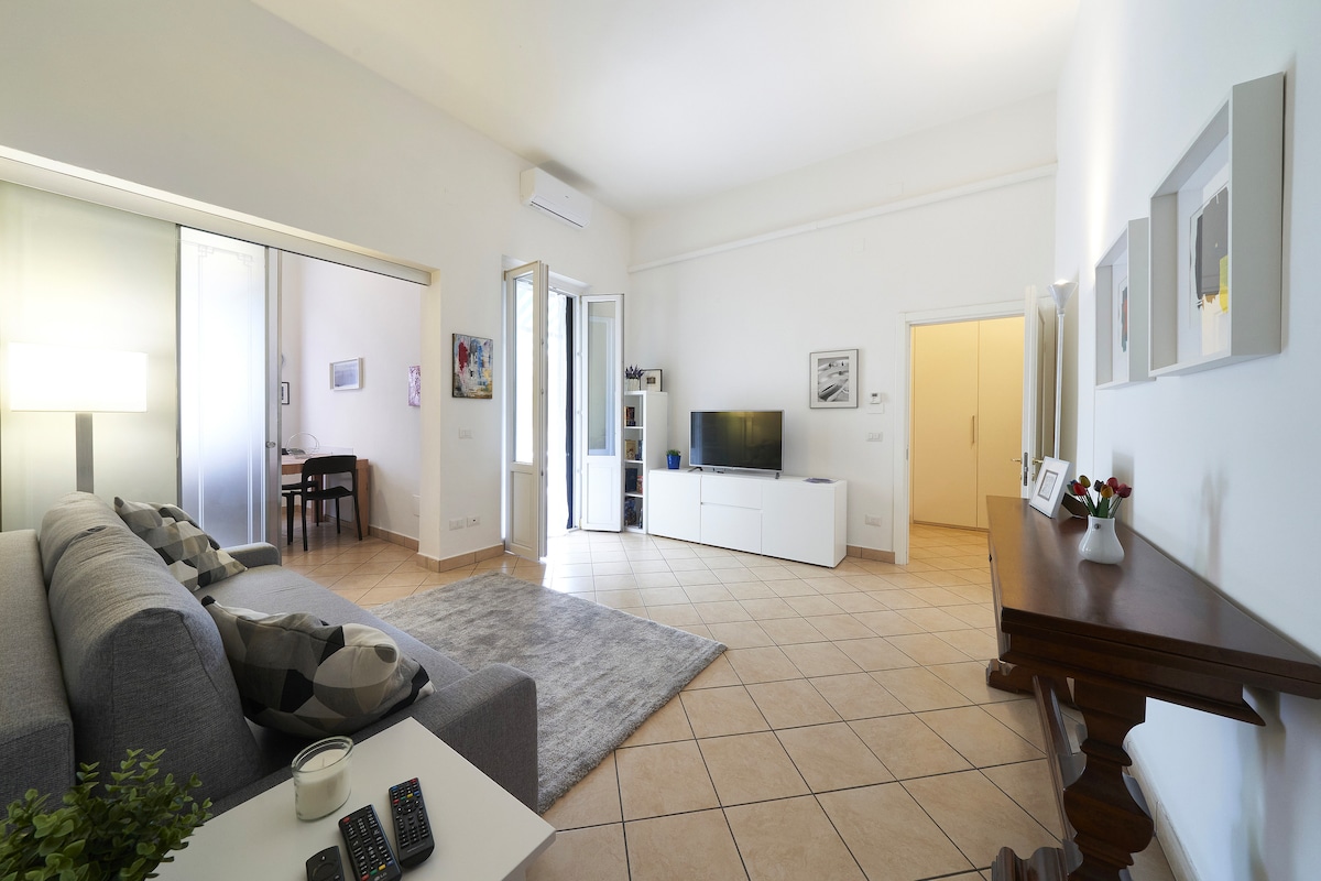 A spacious living area features a comfortable gray sofa, a white television stand, and a flat-screen TV. Light-colored walls reflect natural light from a window, creating a welcoming atmosphere. A small dining table is visible in the background, enhancing the functional layout.