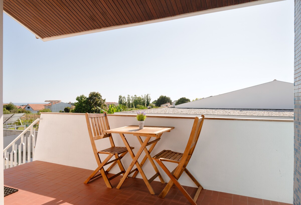 A private balcony is presented, featuring a small wooden table with two folding chairs. A potted plant adds a touch of greenery, while the surrounding area includes rooftops and trees, with clear blue skies above.