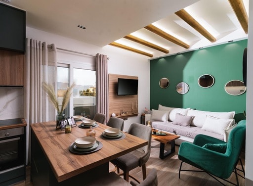 A stylish and bright living area showcases a wooden dining table set for four, complemented by a vibrant green accent wall. A comfortable sofa is positioned against the wall, with circular mirrors reflecting light. Natural light filters in through the window, enhancing the space.