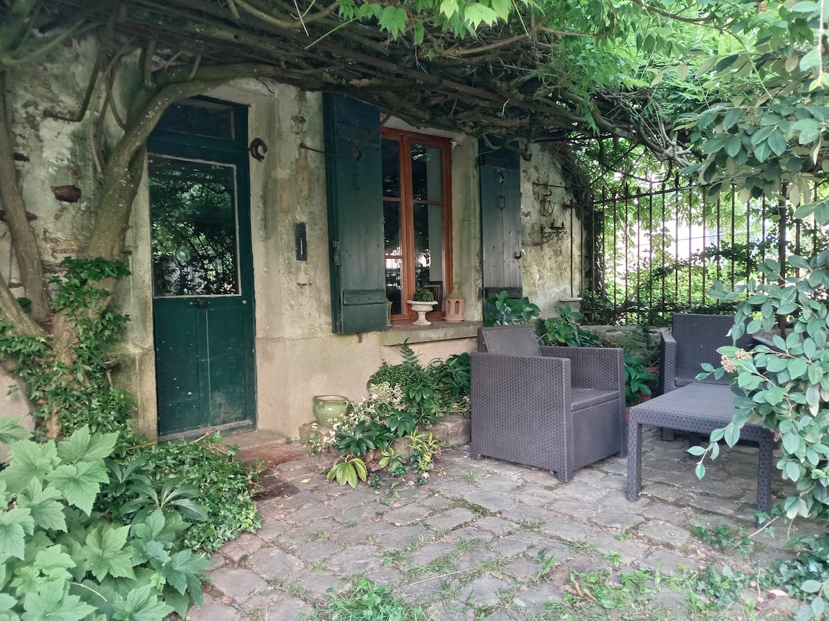 A charming outdoor terrace is framed by lush greenery, featuring a small paved entryway. Two dark wicker chairs are positioned around a small table. A door with green shutters stands adjacent, inviting guests into the serene retreat.