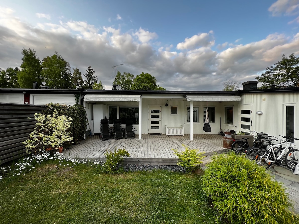 A spacious outdoor deck extends from the house, surrounded by greenery and flower beds. Several bicycles are parked nearby, and a dining area with chairs is visible. The sky above features scattered clouds, highlighting the inviting atmosphere of the outdoor space.