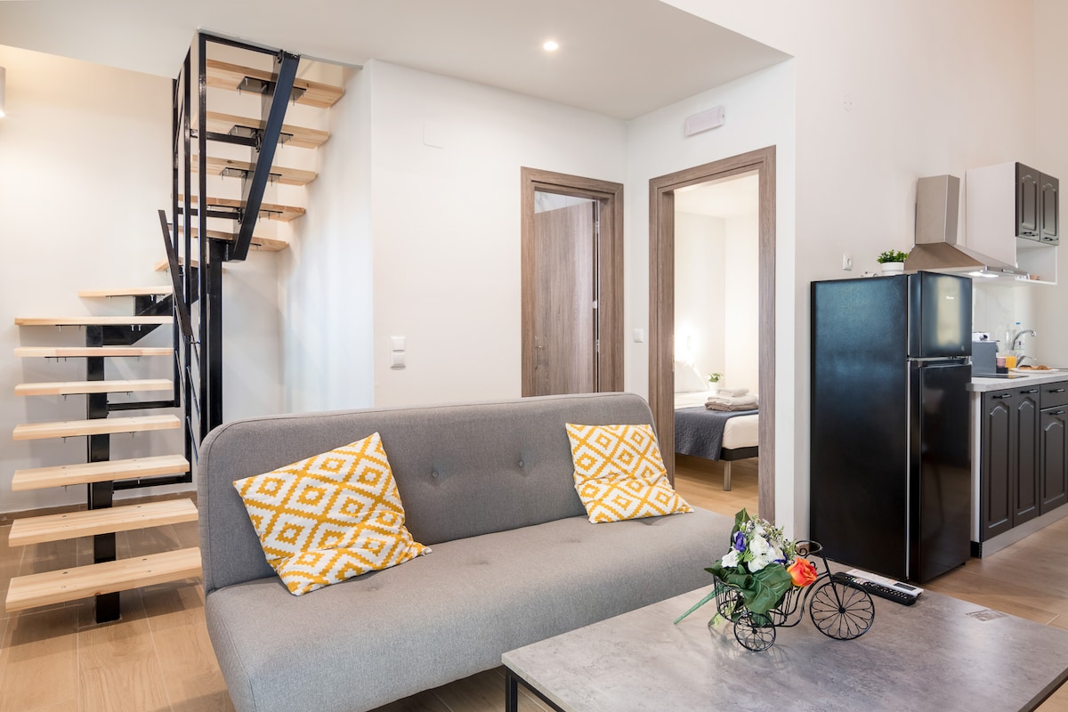 The living area features a comfortable grey sofa with decorative yellow pillows, positioned in front of a minimalist coffee table. A stylish wooden staircase leads to the upper level. The open space includes a kitchenette with a black fridge visible in the background.