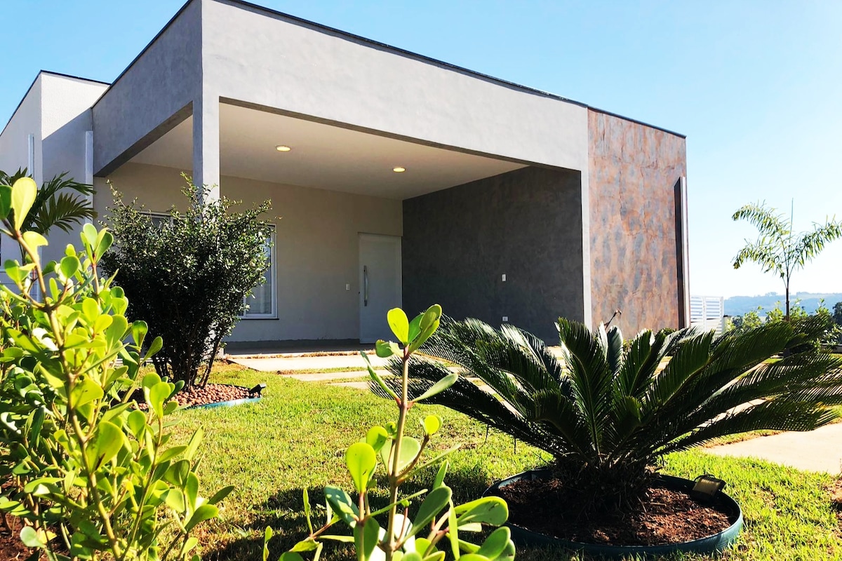The exterior of the house features a modern design with a spacious entrance. Lush greenery surrounds the property, including shrubs and a palm plant, complementing the clean lines of the building. Natural light enhances the inviting facade under a clear blue sky.