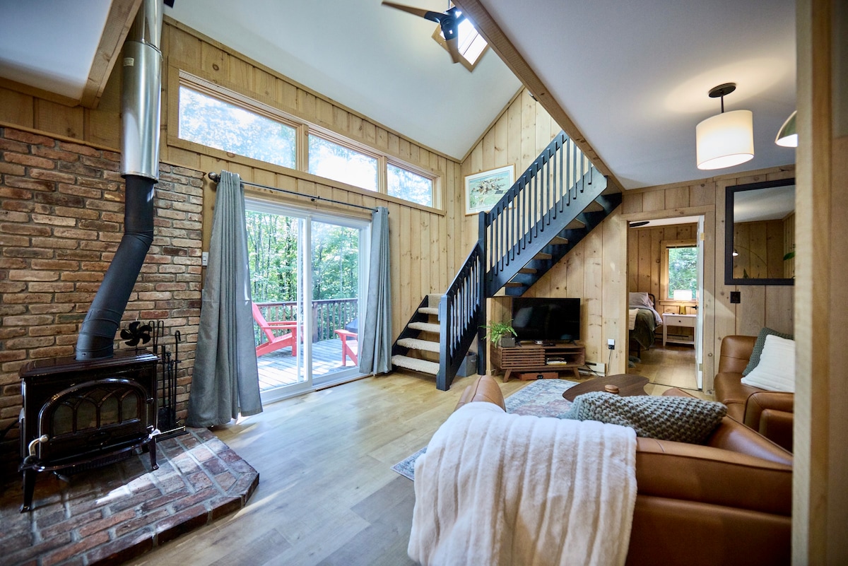 A warm living area features a wood-burning stove against a brick wall, complemented by leather furniture. Large windows provide views of the surrounding greenery, while stairs lead to the upper level. Soft curtains frame the windows, enhancing the cozy ambiance.
