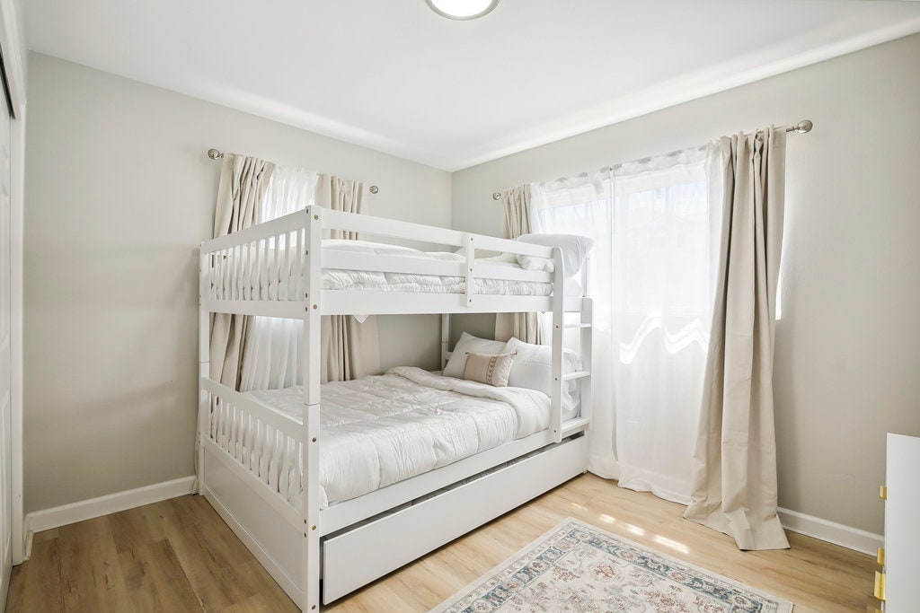 A well-lit bedroom features a white bunk bed with a spacious lower sleeping area. Natural light filters through sheer curtains, highlighting the soft, neutral tones of the decor. A decorative area rug adds warmth to the wooden floor.
