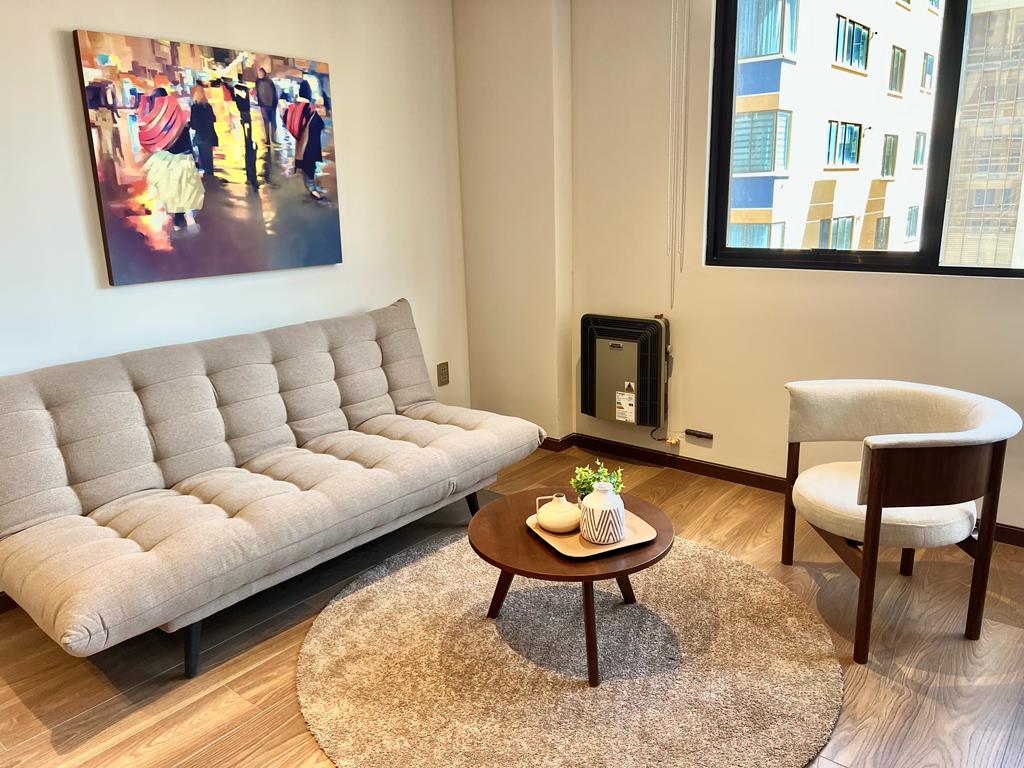 A comfortable seating area features a modern beige sofa bed, a round coffee table, and a light-colored chair. A piece of art depicting vibrant street scenes adorns the wall, while natural light filters in through the window, illuminating the wooden flooring.
