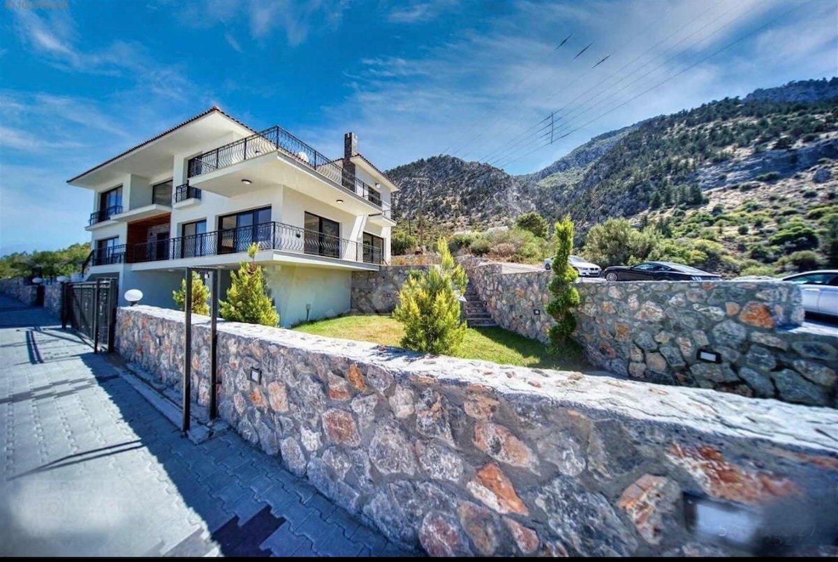 The exterior of the villa is shown, featuring a three-story structure that is embraced by a stone wall. Lush greenery is visible around the property, with a backdrop of rolling hills and a clear blue sky.