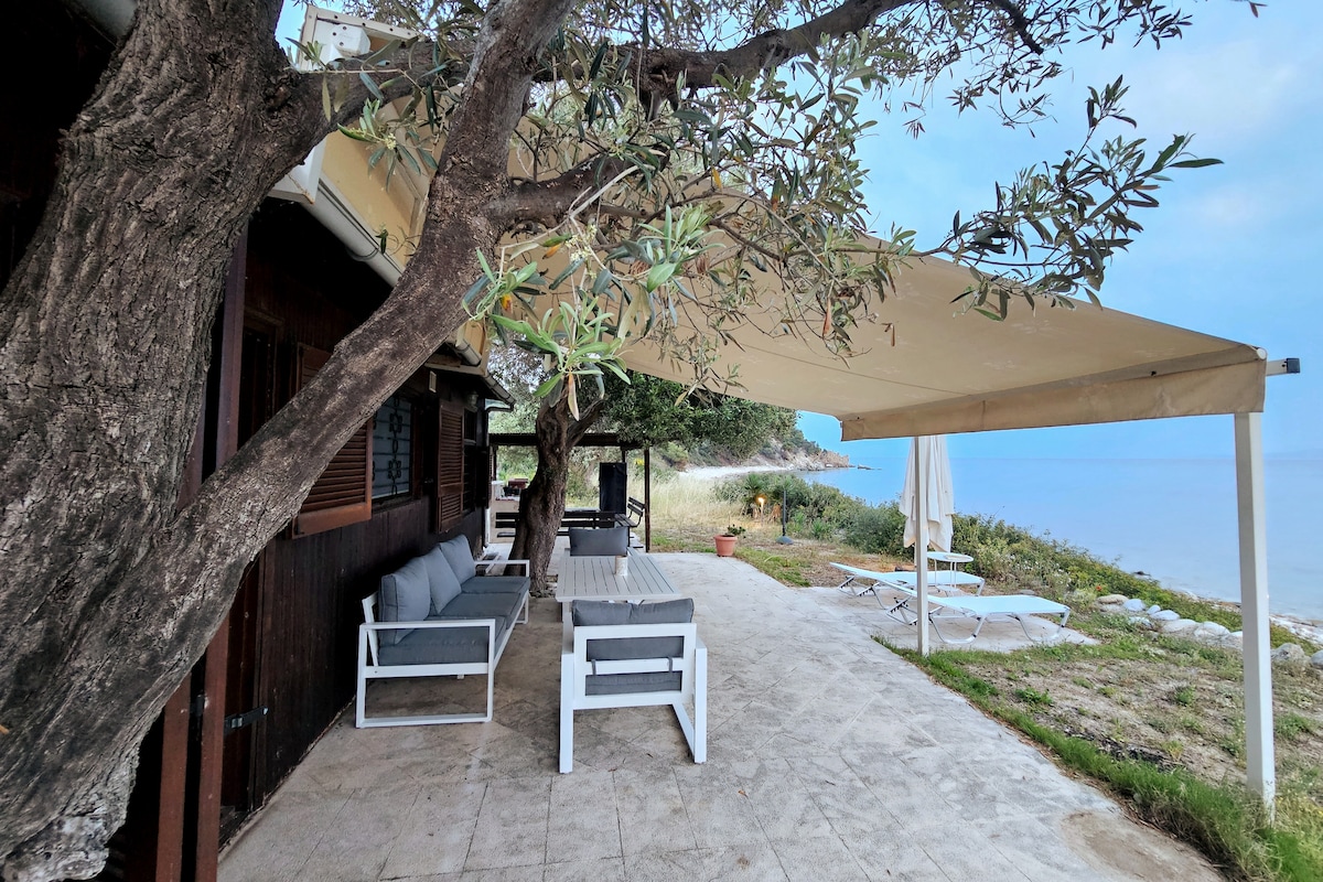 A shaded terrace is shown, featuring a comfortable seating area with white outdoor furniture. The space is surrounded by greenery, with views of the sea in the background. Sunbeds are positioned nearby, inviting relaxation in the gentle coastal breeze.