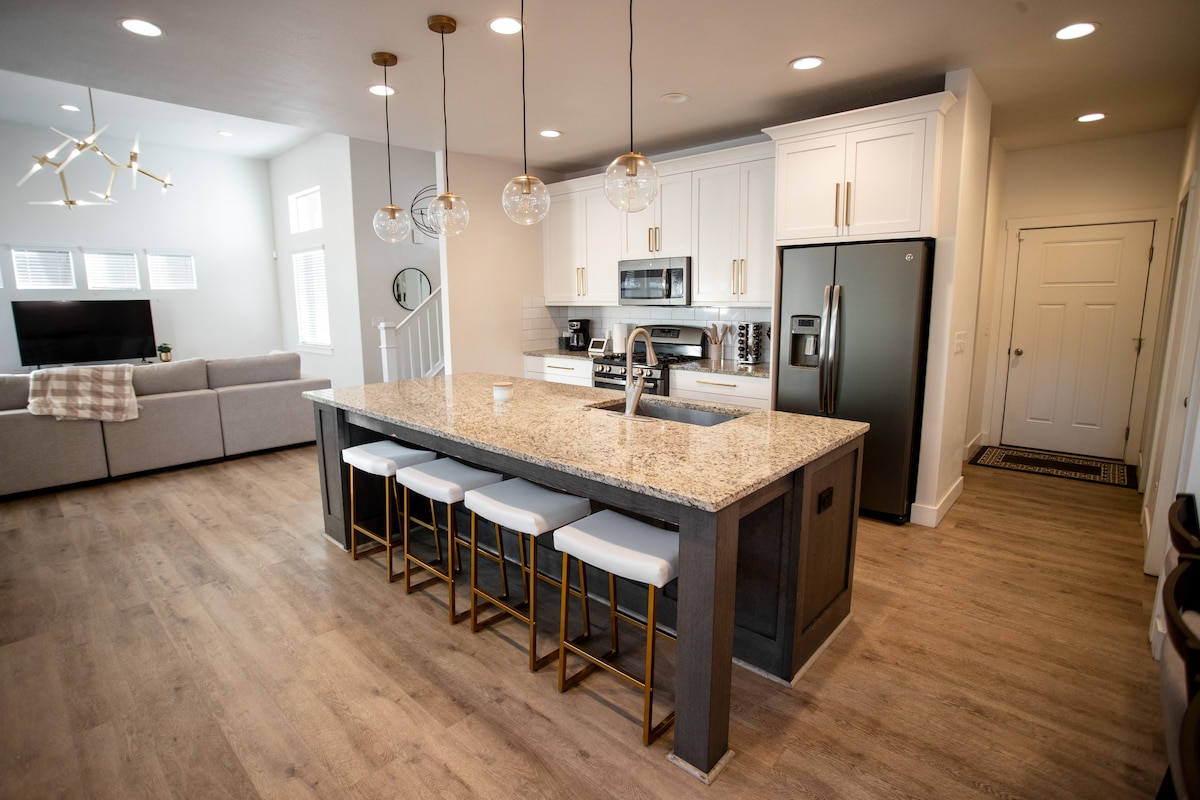 The open kitchen features a large island with seating for four. Modern appliances, including a stove and refrigerator, are positioned conveniently. The adjoining living area is visible, showcasing a comfortable sofa and a decorative light fixture above the island.