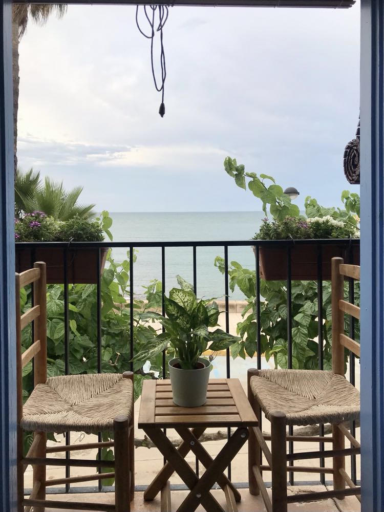 A small balcony is framed by greenery, featuring two wooden chairs and a compact table. A potted plant sits at the center, while the ocean can be seen in the background, enhancing the view with gentle waves and a cloudy sky.