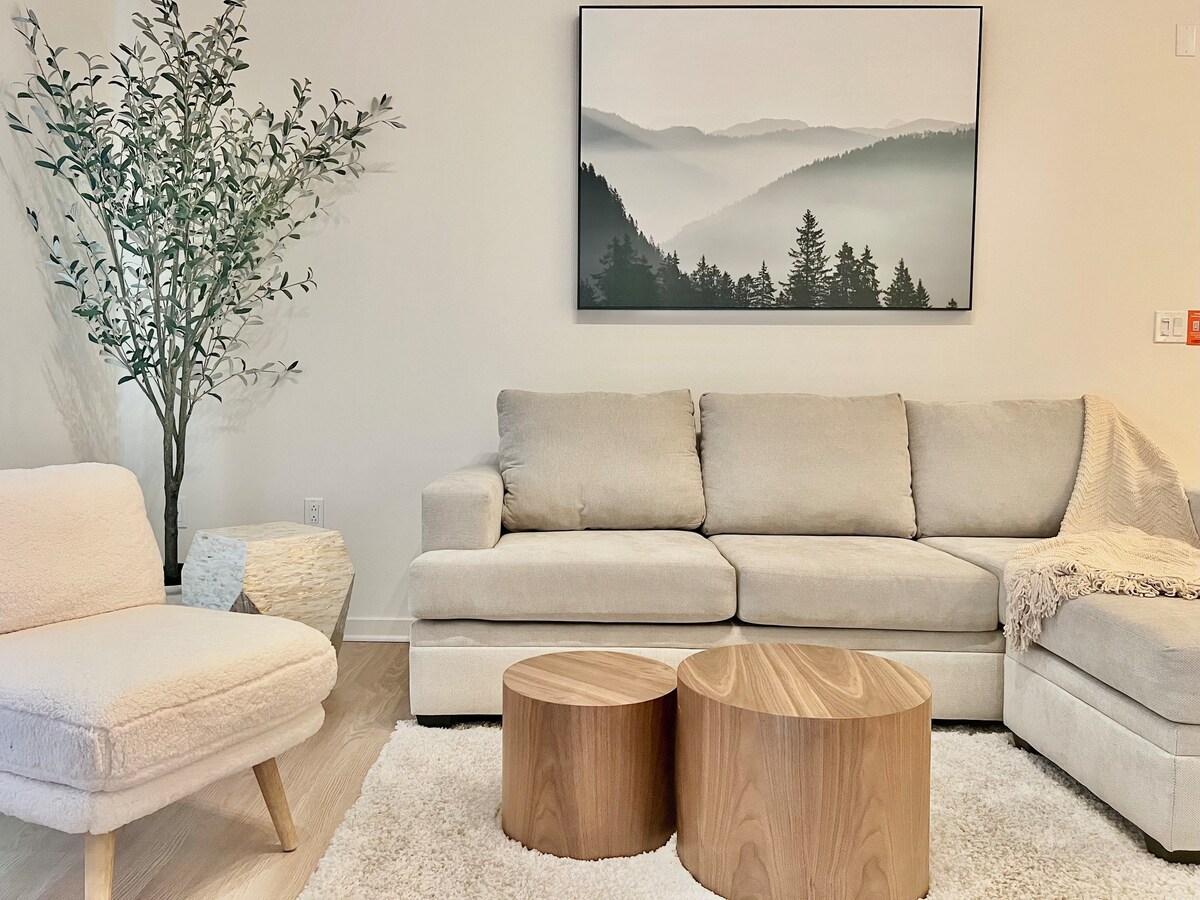 A modern living space features a light-colored sectional sofa complemented by a cozy throw. Natural wood accent tables sit on a soft area rug, creating a comfortable atmosphere. A peaceful landscape painting with soft hues hangs above the sofa, enhancing the room's serene vibe.