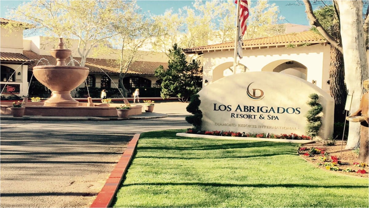 The entrance to Los Abrigados Resort & Spa features a decorative sign surrounded by well-maintained green grass and blooming flowers. A stone fountain is visible in the background, complementing the resort's welcoming atmosphere.