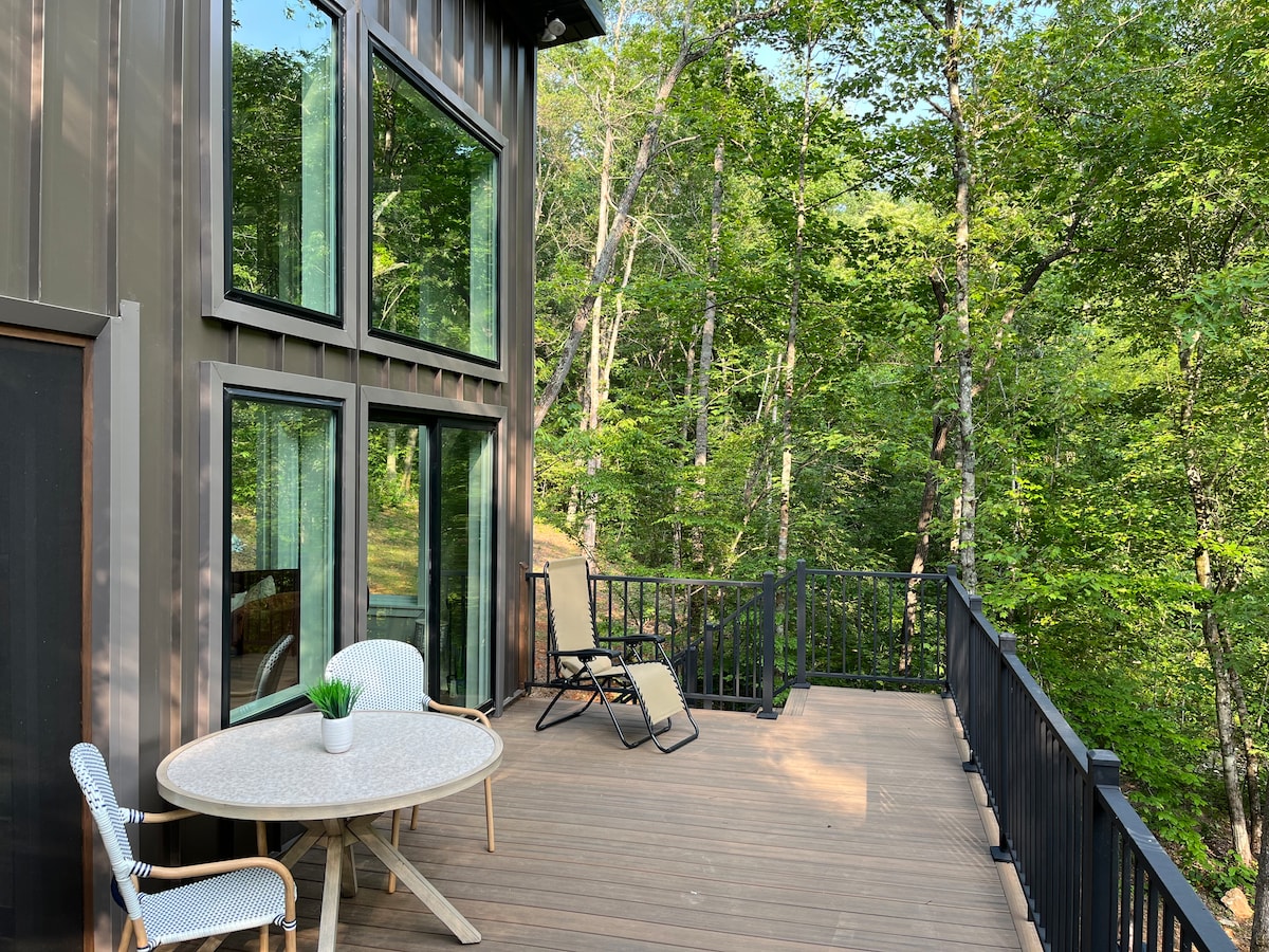 An outdoor deck features a round table with a small potted plant and two lounge chairs. Large glass windows frame the space, offering views of the surrounding trees, creating a seamless connection to nature.