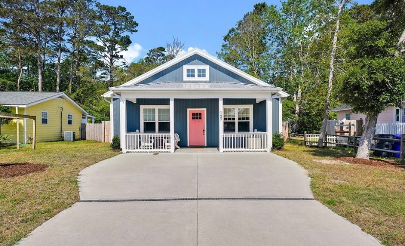 The front view of a modern beach cottage is presented, featuring a welcoming porch with a pink door. Surrounding trees offer shade, while the driveway is neatly paved, leading up to the entrance. The exterior is painted in calming blue tones, complemented by white trim.