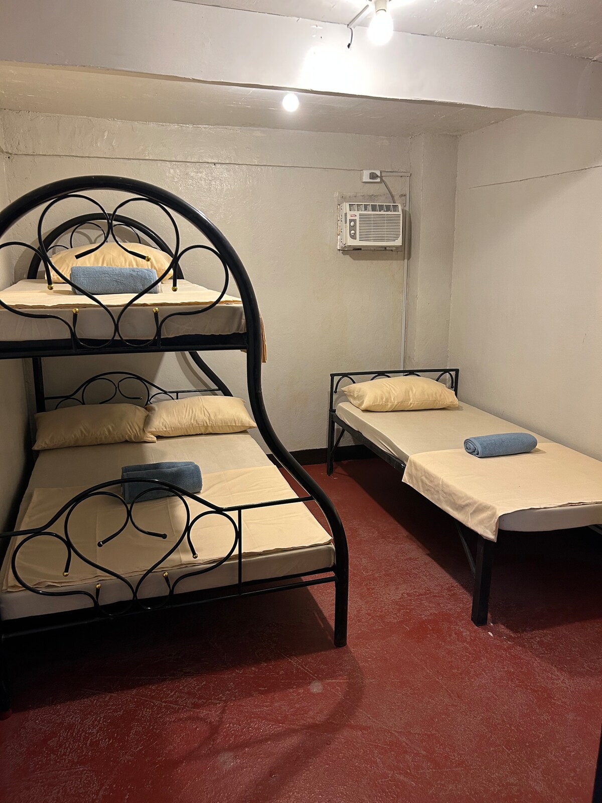 A room is furnished with a bunk bed and a single bed, each topped with neutral-colored linens. Two blue towels are neatly placed on the beds. The space features a red floor and is illuminated by ceiling lights.