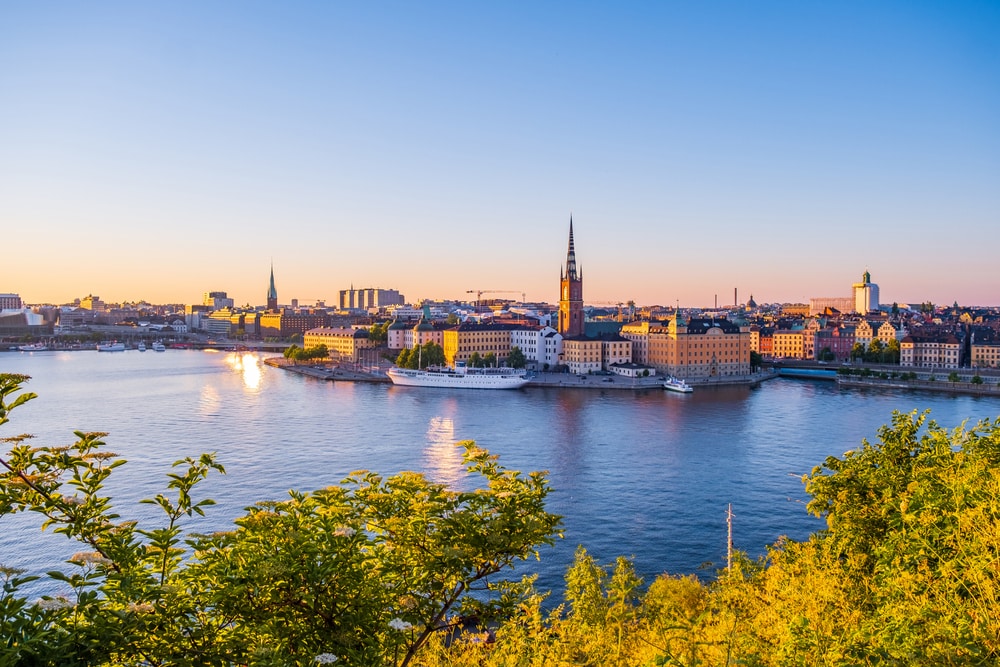 A panoramic view captures the skyline of Stockholm during sunset, with the reflection of light shimmering on the calm water. Historical buildings and modern structures line the waterfront, while greenery in the foreground adds a natural contrast to the cityscape.