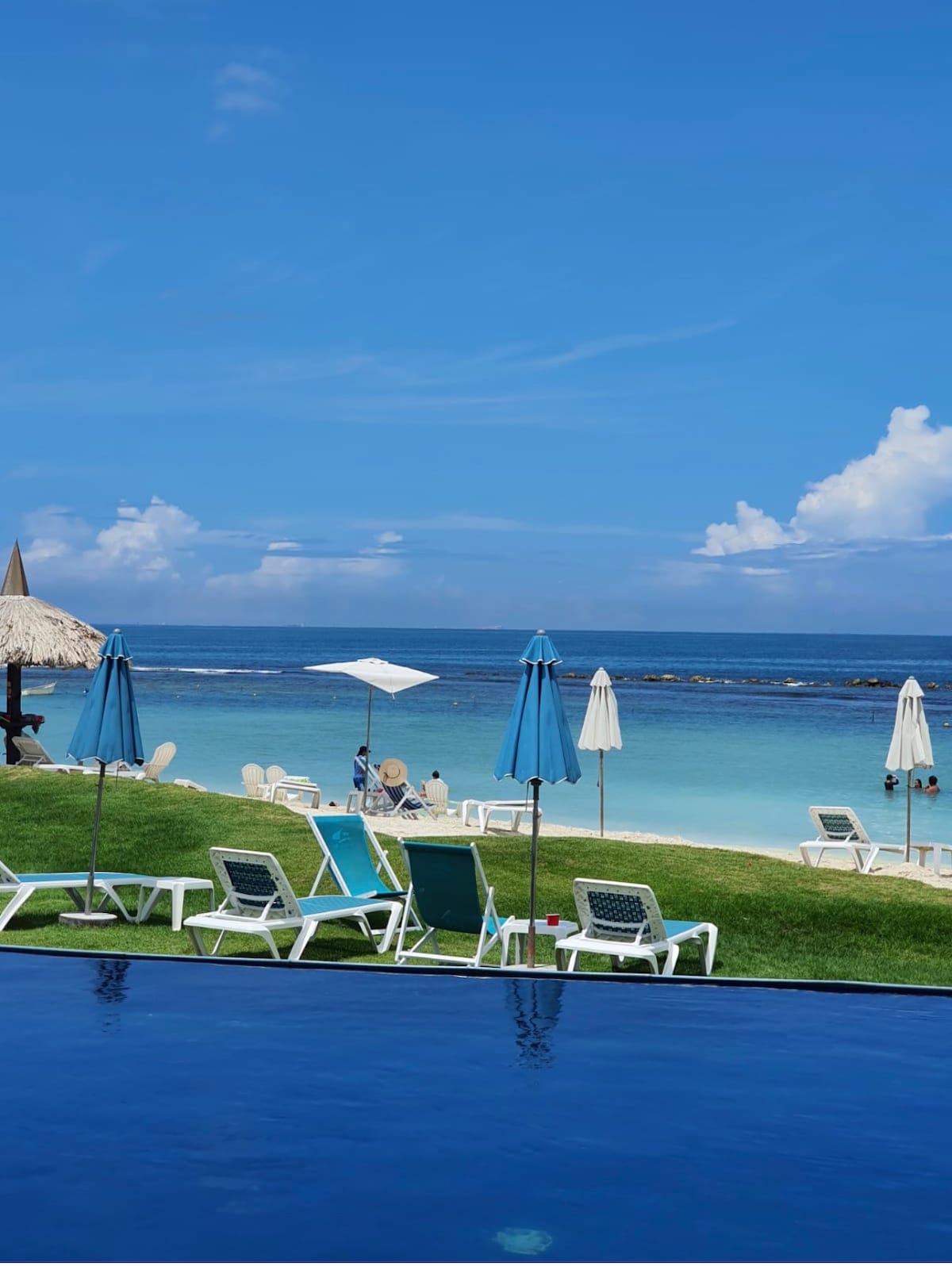 A serene beachfront scene features a smooth sandy shore meeting calm blue waters. Sun loungers and umbrellas are arranged on the grass, overlooking the ocean. A pool edge is visible in the foreground, reflecting the clear sky above with scattered clouds.