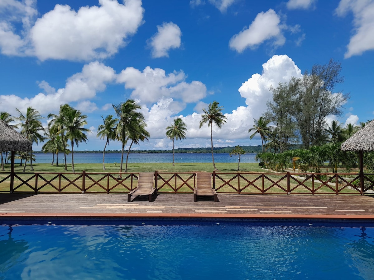 A tranquil outdoor area features a swimming pool surrounded by a wooden deck. Two loungers are positioned for relaxation, with palm trees lining the background. The serene bay and a clear blue sky filled with fluffy clouds create a peaceful atmosphere.