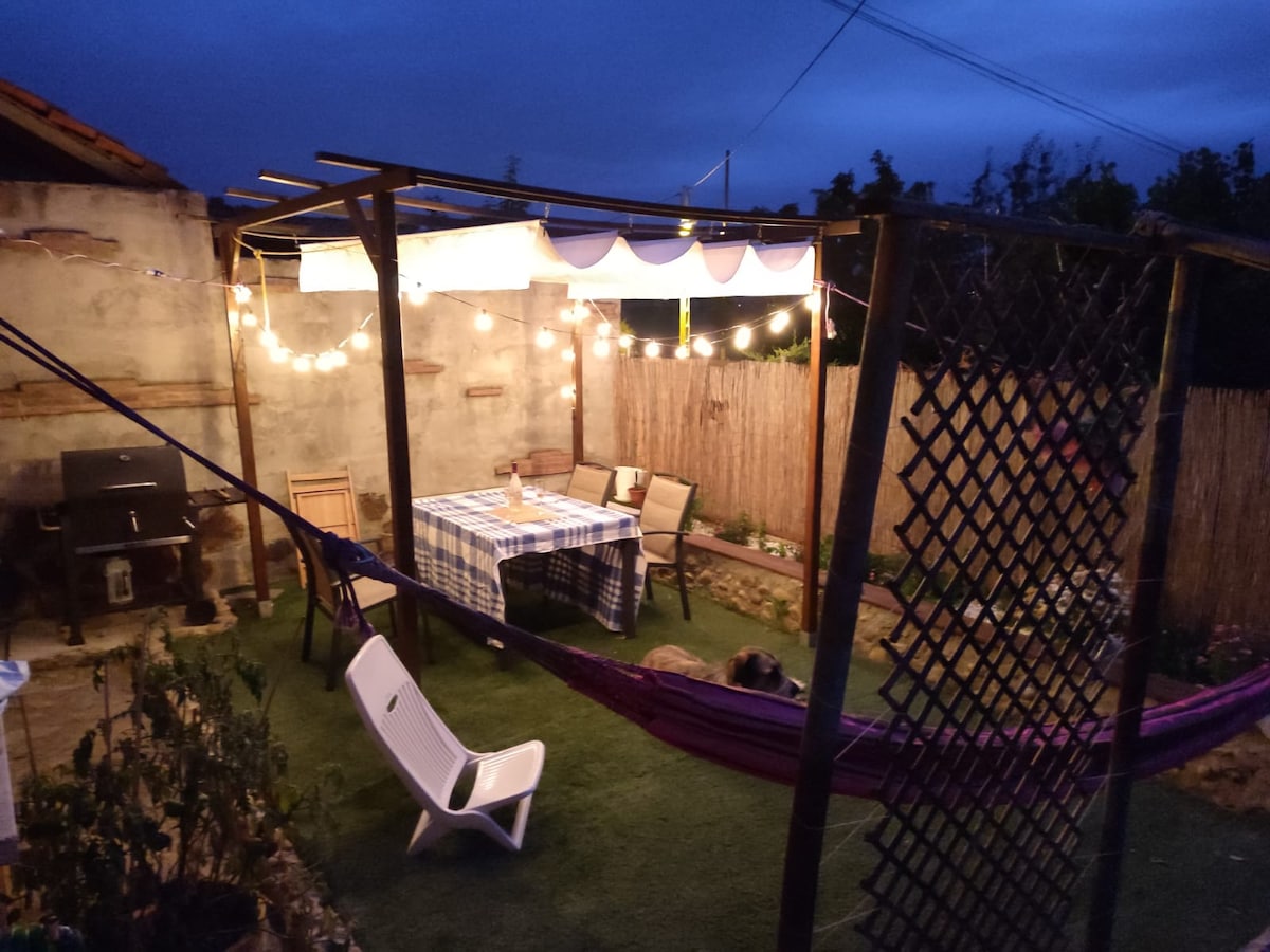 An outdoor patio area is illuminated by string lights, creating a peaceful ambiance. A table is set with checkered cloth and surrounded by chairs. A hammock is hung nearby, and a small barbecue grill is visible in the background, framed by a garden with low greenery.