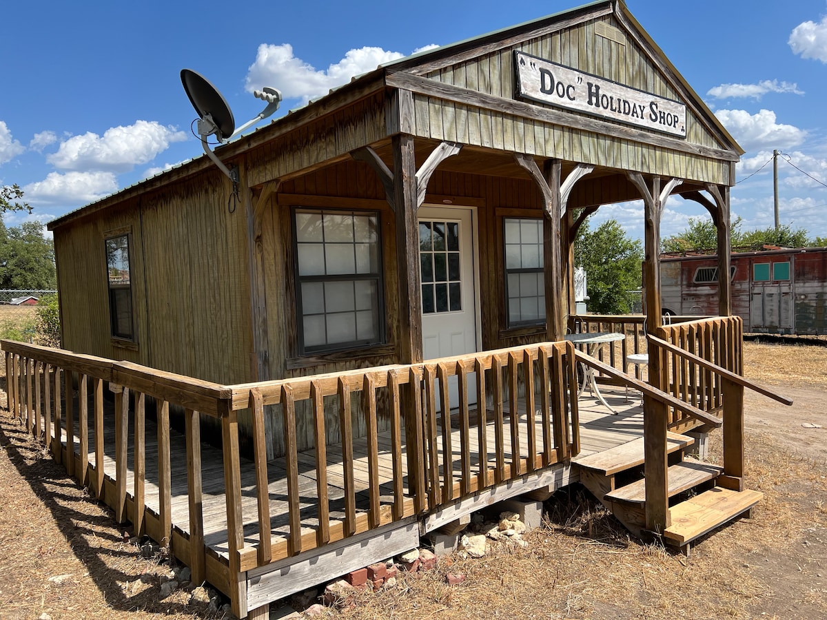The exterior of Doc Holliday Cabin is showcased, featuring wooden siding and a welcoming porch with a railing. The entrance is framed by large windows, and a satellite dish is mounted on the roof. A clear blue sky and fluffy clouds provide a bright backdrop.