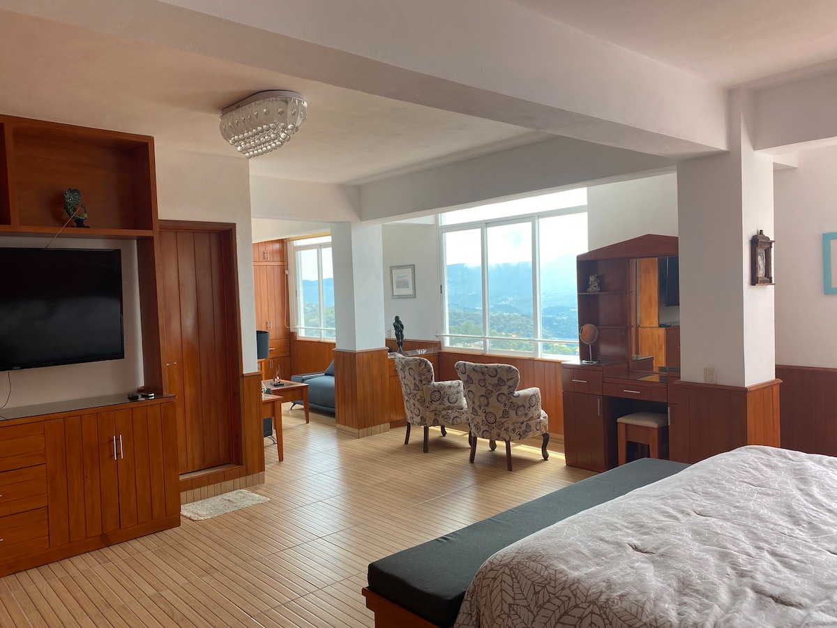 A spacious loft features a large window offering views of the surrounding mountains. Wooden furniture, including a desk and armchairs, creates a comfortable work and relaxation area. A television is mounted on the wall, and light wood flooring adds warmth to the space.